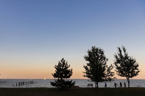 The sun sets over the water at Carr's-Elktonia beach. Three trees, a bench, a dock and a group of people are silhouetted against the sunset.