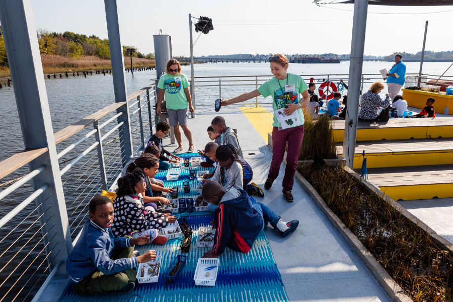 Kids sit on a mat on a barge on the water with two teachers instructing.