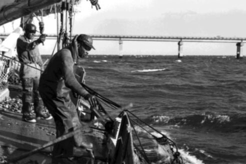 Two African American watermen work on a boat in rough water. They are wearing rubber waders and working rigging and nets.