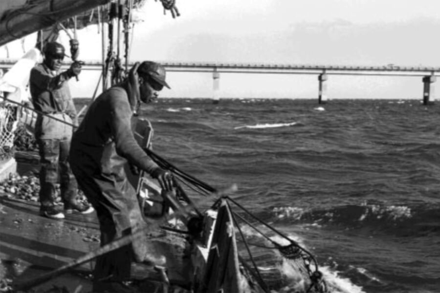 Two African American watermen work on a boat in rough water. They are wearing rubber waders and working rigging and nets.